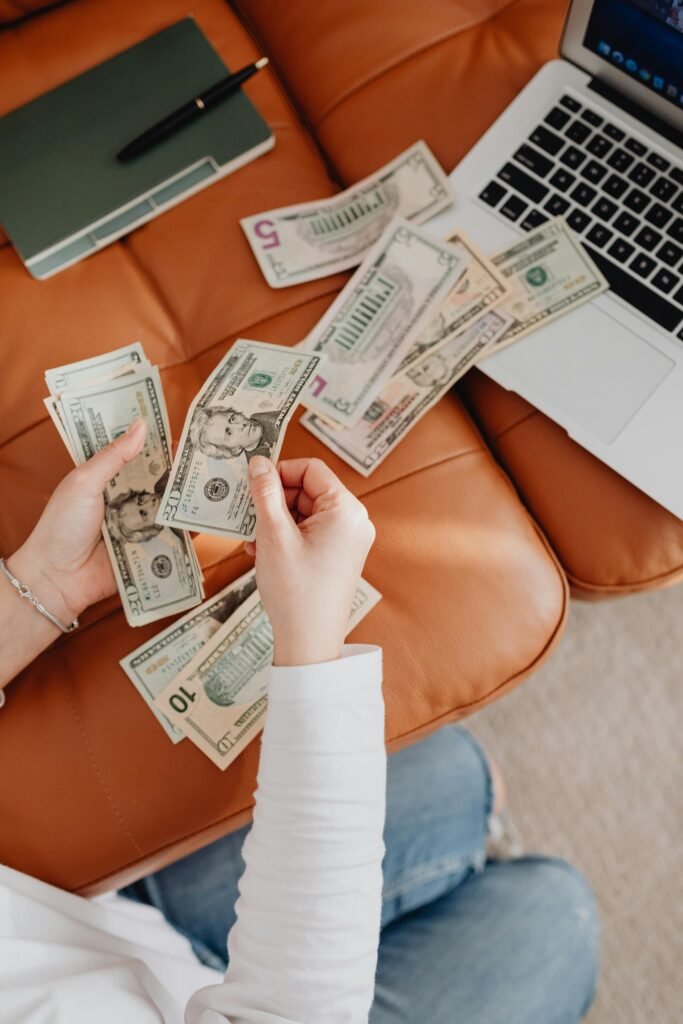 Person counting cash with a laptop and notepad on leather couch, representing finance and budgeting.